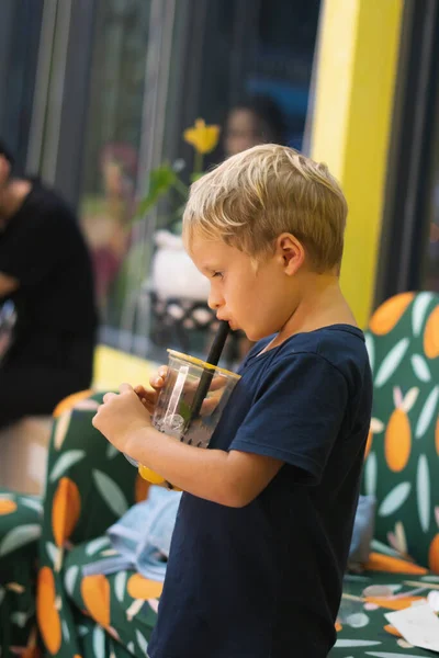 Tampilan samping anak laki-laki pirang yang haus minum limun, teh buah, es teh kombucha dengan persik dan lemon, air susu minuman jeruk dari cangkir plastik dengan jerami, berdiri. Format vertikal cerita — Stok Foto