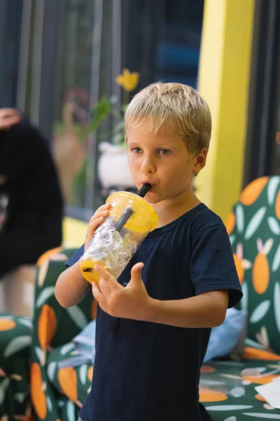 Front view of happy thirsty blond kid boy drinking lemonade, fruit tea, kombucha ice tea with peach and lemon, wellness orange drinks from plastic cup with straw, standing. Stories vertical format — Stock Photo, Image