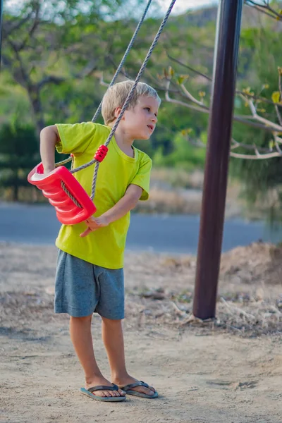 Счастливый блондин маленький мальчик собирается кататься на качелях, играть на детской площадке в парке. Летнее время, детский сад, активность, простые радости детства — стоковое фото
