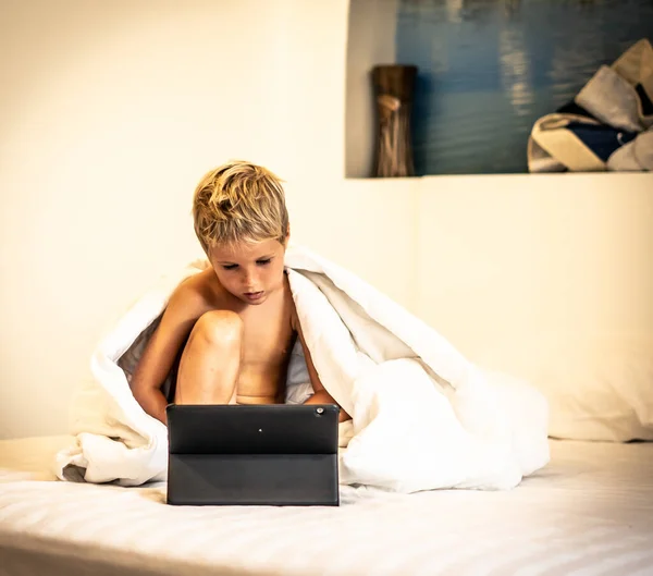 Boy with tablet watch cartoon under blanket in bed in the morning or evening night, when parents busy or sleep. Dependence on TV, difficulties to control childrens content in the Internet concept