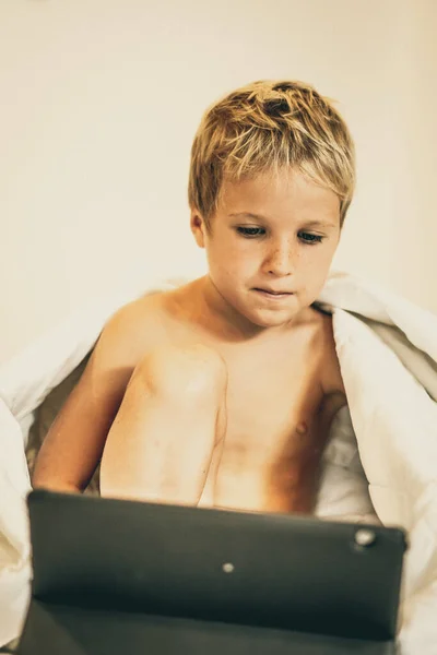 Anak laki-laki dengan tablet menonton kartun di bawah selimut di tempat tidur di pagi atau malam hari, ketika orang tua sibuk atau tidur. Tergantung pada TV, kesulitan untuk mengontrol konten anak-anak dalam konsep Internet — Stok Foto