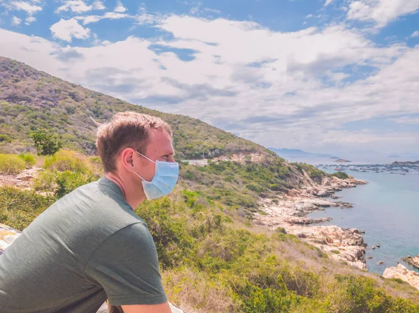 Pemuda tampan melindungi dari kontaminasi udara atau koronavirus dengan memakai topeng medis, berjalan di luar taman dekat laut azure, langit biru. Hentikan infeksi, perlahan-lahan muncul dari pembatasan penguncian — Stok Foto