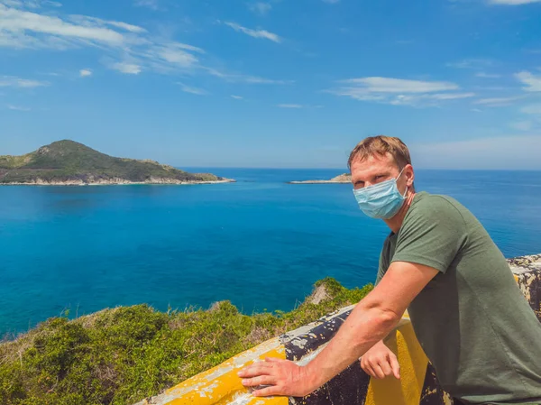 Pemuda tampan melindungi dari kontaminasi udara atau koronavirus dengan memakai topeng medis, berjalan di luar taman dekat laut azure, langit biru. Hentikan infeksi, perlahan-lahan muncul dari pembatasan penguncian — Stok Foto