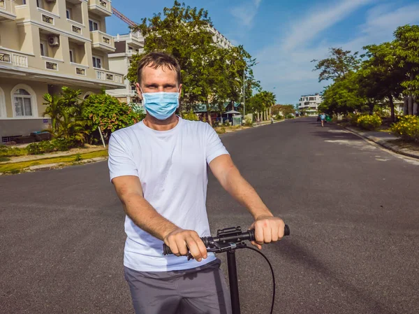 Active outdoor life in surgical sterilizing face mask. Young beautiful man riding on adult scooter in modern cottage village. Protecting from air pollution and virus infection as new normal lifestyle