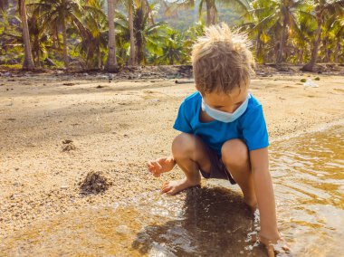 Kumsalda kumla oynayan, tıbbi maske takan sevimli sarışın çocuk. Hava kirliliğinden ve virüs enfeksiyonundan korunarak kovid19 virüs salgını sırasında yeni normal yaşam tarzı olarak