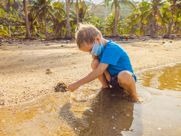 समुद्र तट पर मेडिकल फेस मास्क के साथ प्यारा छोटा युवा गोरा लड़का रेत के साथ खेल रहा है। कोविद 19 वायरस महामारी युग के दौरान नई सामान्य जीवन शैली के रूप में वायु प्रदूषण और वायरस संक्रमण से रक्षा करना — स्टॉक फ़ोटो, इमेज