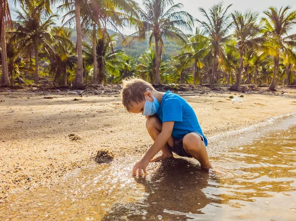 해변에서 의학용 마스크를 쓰고 모래를 가지고 노는 귀여운 금발의 소년. 공습 범바이러스 유행 시대의 새로운 일상 생활 방식으로 대기 오염과 바이러스 감염으로부터 보호하는 일 — 스톡 사진