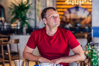 Ciddi sarışın, sıradan tarzda kırmızı tişörtlü bir adam modern kafede oturuyor, gergin bir şekilde birini bekliyor ya da değiştiriyor, düşünüyor. Deri küçük photoshop. İnsani ilişkiler, covid sırasında yaşanan zorluklar ya da yaşam sorunları