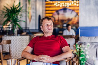 Ciddi sarışın, sıradan tarzda kırmızı tişörtlü bir adam modern kafede oturuyor, gergin bir şekilde birini bekliyor ya da değiştiriyor, düşünüyor. Deri küçük photoshop. İnsani ilişkiler, covid sırasında yaşanan zorluklar ya da yaşam sorunları