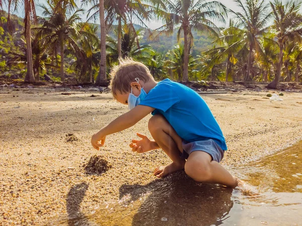 Χαριτωμένο ξανθό αγοράκι με μάσκα προσώπου στην παραλία που παίζει με άμμο. Προστασία από την ατμοσφαιρική ρύπανση και τη μόλυνση από ιούς ως νέο φυσιολογικό τρόπο ζωής κατά την εποχή της επιδημίας του ιού covid19 — Φωτογραφία Αρχείου