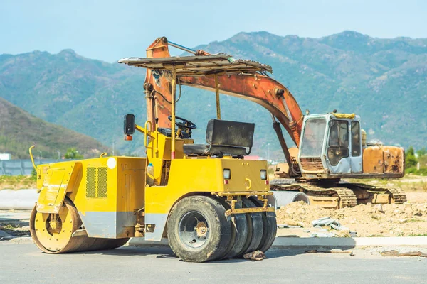 Big yellow road rollers and other construction machinery in sunny day work on new modern district road construction site under blue sky and mountain background. Civil building industry concept