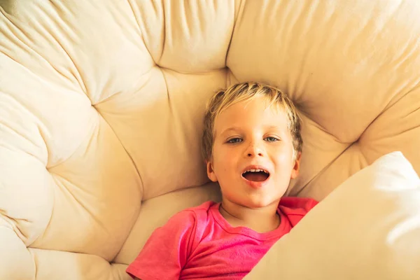 Blond skrattar med rolig pojke öppna munnen i rosa t-shirt sitta med kudde i rund stol inomhus. Familjeförhållande, lycklig barndom, problembeteende, utbildningspsykologi — Stockfoto