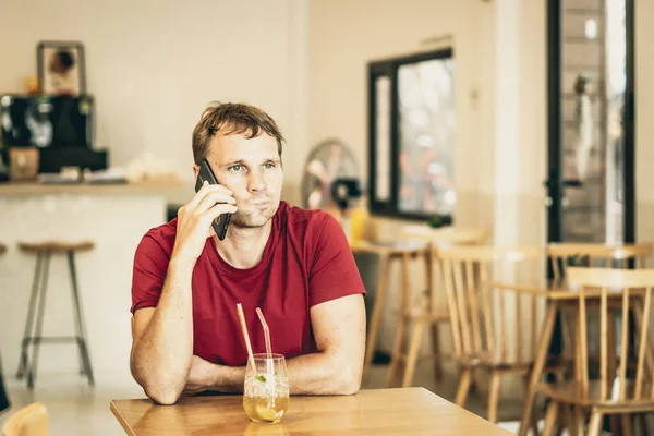Serious sad young man answering smart phone in red shirt in cafe drinking ice tea, waiting somebody or coworking. Real people expression, life problems solving or communication, relationship