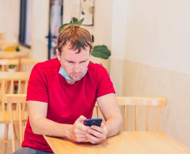 Tıbbi maske takan, ciddi ciddi düşünceli, düşünceli bir genç kafedeki kırmızı tişörtlü telefonla mesaj gönderiyor. Gerçek insan ifadesi, hayat sorunları çözme, Covid salgını sırasında iletişim.