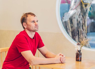 Gülümse, kırmızı tişörtlü yakışıklı adam modern kafede oturuyor, mutlu bakışlar atıyor, birini bekliyor ya da tanışıyorlar. Gerçek insanların ifadesi, insan ilişkileri, iyi gelecek konsepti değiştirir.