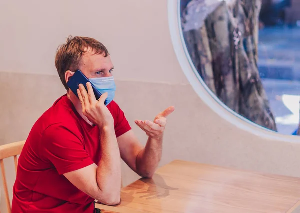 Young serious pensive attentive mean in medical face mask tense thinking speak answer phone in red t-shirt in cafe. Real people expression, life problems solving, communication during Covid Pandemic
