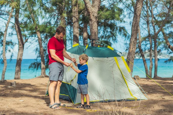 Camping orang asing gaya hidup wisatawan memasang mendirikan berkemah hijau abu-abu mereka di hutan musim panas dekat laut lazur. Anak laki-laki yang lucu membantu ayahnya merakit tenda. Pendidikan alami anak-anak — Stok Foto