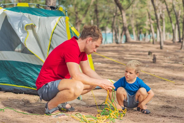 Camping people outdoor lifestyle tourists putting up setting up their green grey campsite in summer forest near lazur sea. Cute boy son helps his father assembling tent. Natural education of children