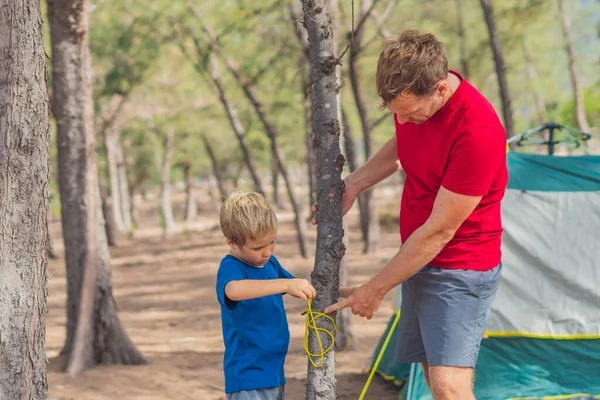 Camping people tourists putting up setting up green grey campsite summer forest near lazur sea. Natural wild live education of children. Father shows tying rope around tree, son helps assembling tent
