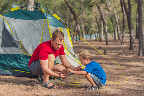 Camping people outdoor lifestyle tourists putting up setting up green grey campsite summer forest near lazur sea. Boy son helps father assembling tent, study metal peg. Natural education of children