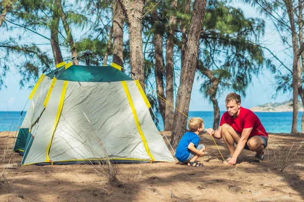 Camping people outdoor lifestyle tourists put up set up green grey campsite summer forest near lazur sea. Boy son helps father study mechanism of modern easy to fold tent. Natural children education