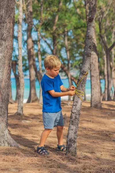 Camping orang asing gaya hidup wisatawan di hutan musim panas dekat laut lazur. Anak laki-laki gemuk serius dalam biru t-shirt belajar teknik bertahan hidup, metode praktek mengikat tali knot. Pendidikan anak alami — Stok Foto