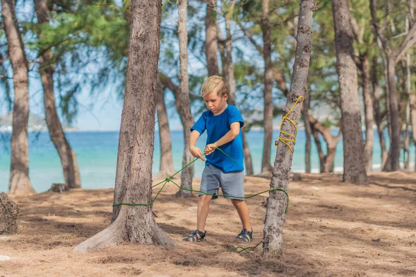 Camping människor utomhus livsstil turister i sommarskogen nära lazur havet. Blond seriös pojke i blå t-shirt studera överlevnadsteknik, metoder för att binda rep knutar. Naturlig utbildning för barn — Stockfoto