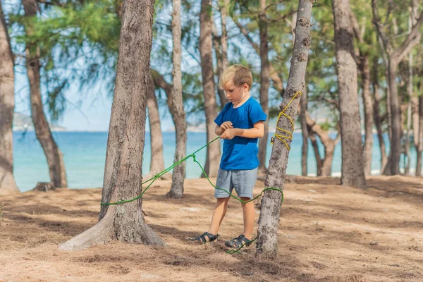 ラズール海の近くの夏の森の中で人々のアウトドアライフスタイル観光客をキャンプ。青いTシャツのブロンドの深刻な少年は、生存技術、ロープノットを結ぶ練習方法を研究しています。自然児教育 — ストック写真