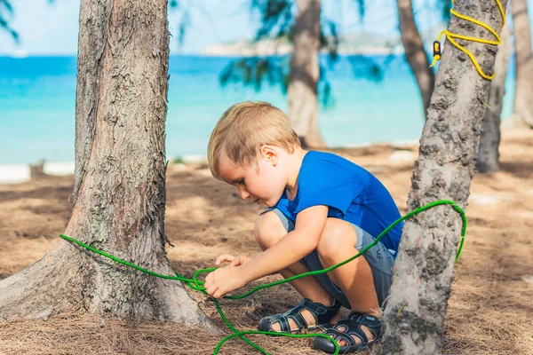 Camping människor utomhus livsstil turister i sommarskogen nära lazur havet. Blond seriös pojke i blå t-shirt studera överlevnadsteknik, metoder för att binda rep knutar. Naturlig utbildning för barn — Stockfoto
