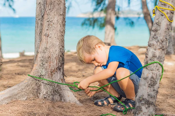 Κάμπινγκ άνθρωποι υπαίθρια τουρίστες τρόπο ζωής στο δάσος καλοκαίρι κοντά στη θάλασσα lazur. Ξανθό σοβαρό αγόρι σε μπλε t-shirt μελέτη τεχνικές επιβίωσης μελέτη, πρακτική των μεθόδων πρόσδεσης κόμπους σχοινί. Φυσική αγωγή των παιδιών — Φωτογραφία Αρχείου