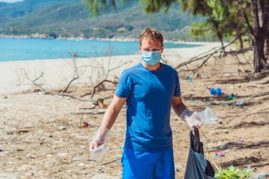 Yüz maskeli gönüllü bir adam deniz kenarındaki çöpleri topluyor, atılan plastik bardaklara bakıyor. Dökülen çöp sorunu, gezegen kirliliği çevre koruma kavramı