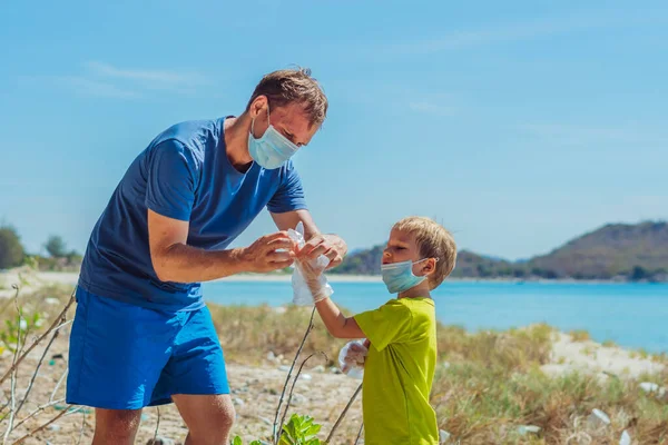 Sukarelawan bertopeng wajah medis biru. Ayah menempatkan sarung tangan pada anaknya untuk mengambil sampah yang mencemari pantai dekat laut. Masalah tumpah sampah planet polusi konsep perlindungan lingkungan — Stok Foto