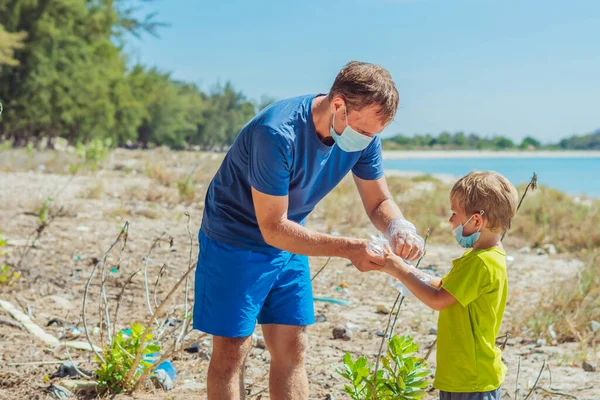 Volunteers in blue medical face mask. Father puts gloves on his son for pick up garbage which pollute beach near sea. Problem of spilled rubbish trash planet pollution environmental protection concept — Stock Photo, Image