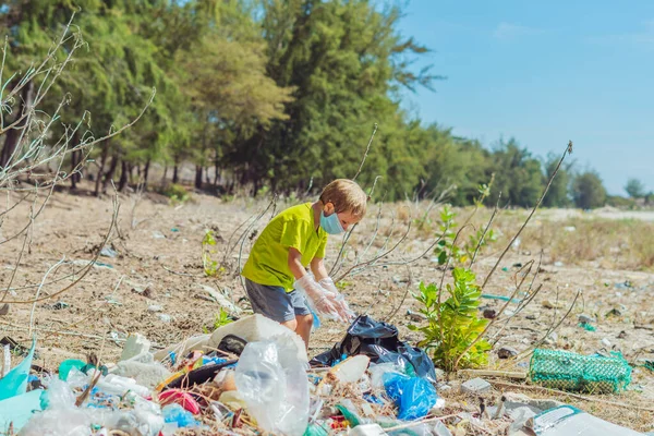 Volunteer boy in face mask helps to pick up garbage which pollute beach near forest. Problem of spilled rubbish trash planet pollution environmental protection concept. Natural children education