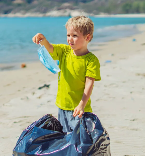 Vrijwilligers strand zand lazur zee. Jongen raapt afval op in een zwarte zak. Zoon weigert een blauw gezichtsmasker te dragen omdat hij moe is. Een nieuw tijdperk in masker. Gemorste rommel. Natuurlijke kinderopvoeding — Stockfoto