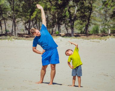 Küçük çocuklar için spor. Yakışıklı baba ve yakışıklı oğul orman parkının yakınındaki kumsalda egzersiz yapıyor, temiz havanın tadını çıkarıyor, doğru çömelme ve yan bükme tekniği uyguluyor.