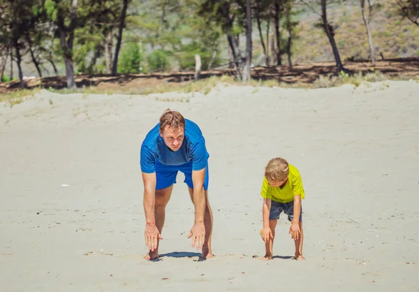 Olahraga untuk anak kecil. Ayah yang tampan dan anak yang lucu berolahraga di pantai pasir dekat taman hutan, memiliki olahraga bersama-sama di udara segar, benar berjongkok dan teknik tikungan ke depan — Stok Foto