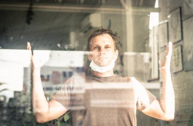 Beyaz koruyucu maske takan bir adam, pencereden dışarı bakıyor, mutlu umut dolu gülümseme gözleri, evdeki hastalığın yakında geçeceğine dair işaretler gösteriyor, COVID 19 Coronavirus salgını nedeniyle kendini izole etme.