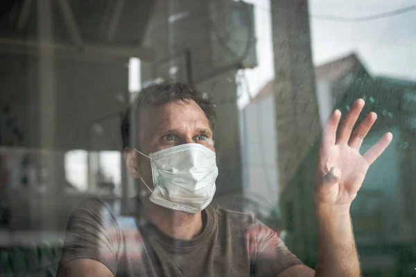 Pria mengenakan topeng pelindung putih, melihat ke luar melalui jendela menyentuh kaca, takut harapan stres di mata, pemulihan dari penyakit di rumah, isolasi diri karena pandemi global COVID 19 Coronavirus — Stok Foto