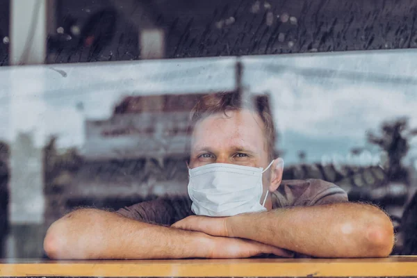 Pria mengenakan topeng pelindung putih, melihat keluar melalui jendela, takut harapan stres di mata, pemulihan dari penyakit di rumah, isolasi diri karena pandemi global COVID 19 Coronavirus — Stok Foto