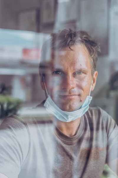 Portrait Man mengenakan topeng pelindung putih, melihat ke luar melalui jendela, harapan yang bahagia mengedipkan mata tersenyum, pemulihan dari penyakit di rumah, isolasi diri karena pandemi global COVID 19 Coronavirus — Stok Foto