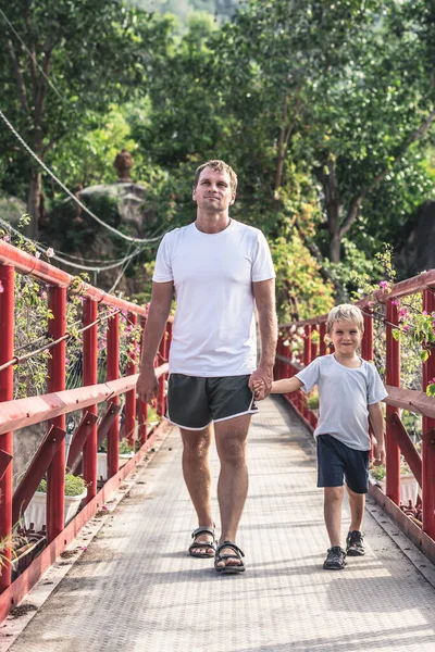 Father son walking across red iron suspension bridge way to forest park. Friendly family vacation outdoor enjoying leisure time nature concept. Dad holds baby by hand, go towards the camera — Stock Photo, Image