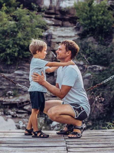 Father hugs son sitting squatting, look each other, smile. Happy childhood, fathers day, mountain, chain background as symbol of strong bond between dad and child. Father influence on son worldview — Stock Photo, Image