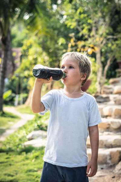 Tampilan depan anak pirang yang haus minum teh hitam dari botol plastik berdiri di luar di latar belakang taman musim panas alam. Konsep minuman sehat dan tidak sehat. Format vertikal cerita — Stok Foto