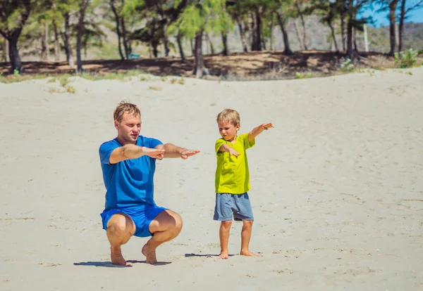 Olahraga untuk anak kecil. Ayah lucu anak berolahraga di pantai pasir dekat taman hutan, memiliki latihan bersama-sama di udara segar, benar jongkok teknik, ayah pengaruh pada anak laki-laki pandangan dunia — Stok Foto