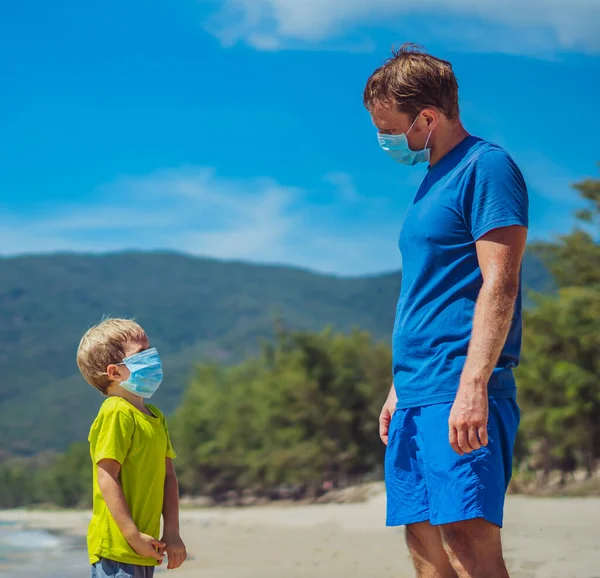 New normal reality safety during COVID 19 Global pandemic. Father and son looking on each other, wearing face blue mask protect coronavirus even walking nature sand sea beach near forest park sun day