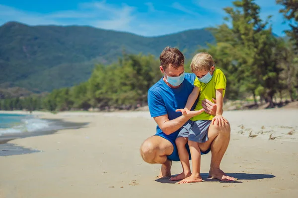 Батько присідає, тримаючи сина, дивлячись на відкриту руку, грають порівняти свої долоні, носячи блакитну маску, захищаючи коронавірус, що йде на природу піщано-морський пляж біля лісового парку сонячний день. COVID нормальна реальність — стокове фото