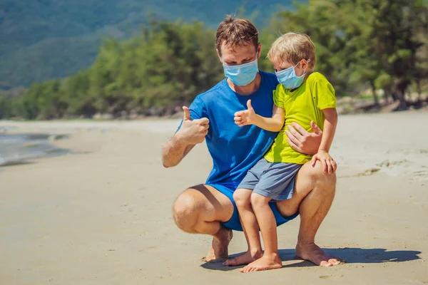 Ayah jongkok memegang anak bermain tinju dengan jempol ke atas, mengenakan topeng biru melindungi coronavirus berjalan alam pasir pantai laut dekat hutan taman matahari hari. Realitas virus pandemi global COVID Stok Gambar