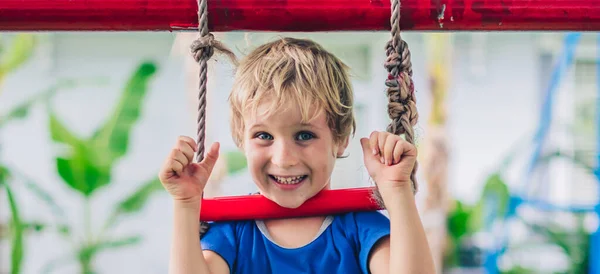 Potret dekat Senyum gembira mata biru bermata biru anak kecil pirang lucu duduk di tangga tali, bermain di luar di taman bermain di TK. penitipan anak, aktivitas, sukacita sederhana masa kecil, musim panas — Stok Foto