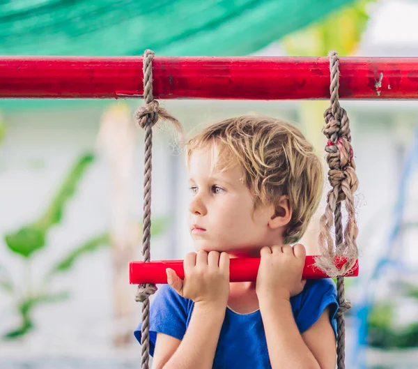 Potret dekat sedih kesepian bintik-bintik biru mata kecil pirang lucu di t-shirt duduk di tangga tali di luar taman bermain di taman kanak-kanak. Penitipan anak, aktivitas, masa kecil, persahabatan, hubungan — Stok Foto